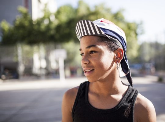 young afro boy with hat and bandana plays basketball on the court of his neighborhood